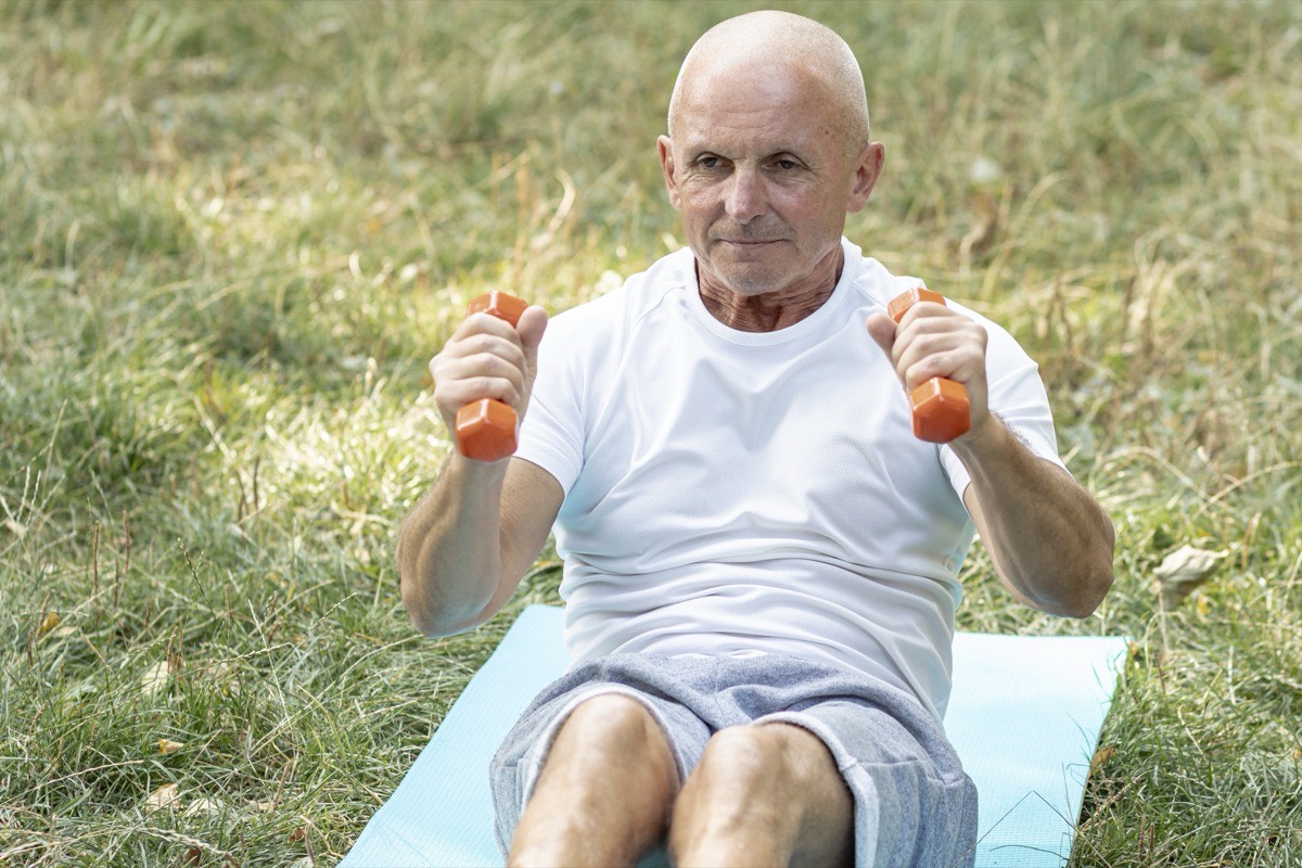 Homme senior réalisant un exercice adapté avec de petits haltères, assis sur un tapis en extérieur